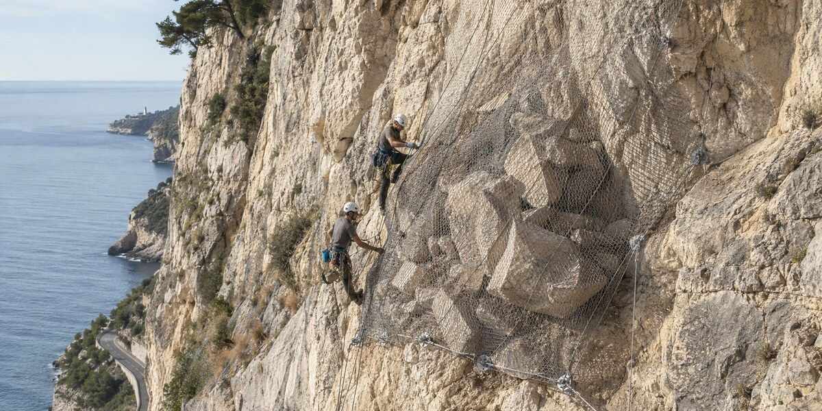 Deux cordistes sécurisant des blocs rocheux derrière un filet métallique sur une falaise méditerranéenne évoquant le Cap de Nice
