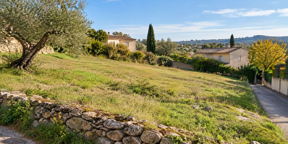 Terrain herbeux en pente douce, bordé d’un muret en pierre, avec une voie d’accès et des maisons en arrière-plan.