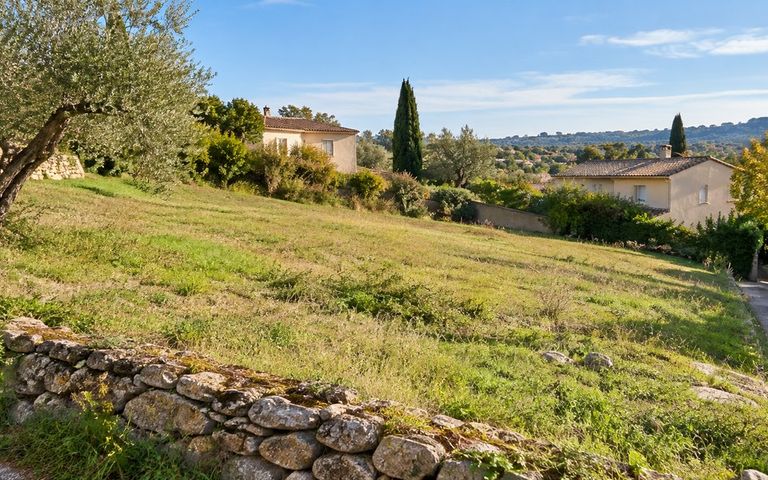 Terrain herbeux en pente douce, bordé d’un muret en pierre, avec une voie d’accès et des maisons en arrière-plan.