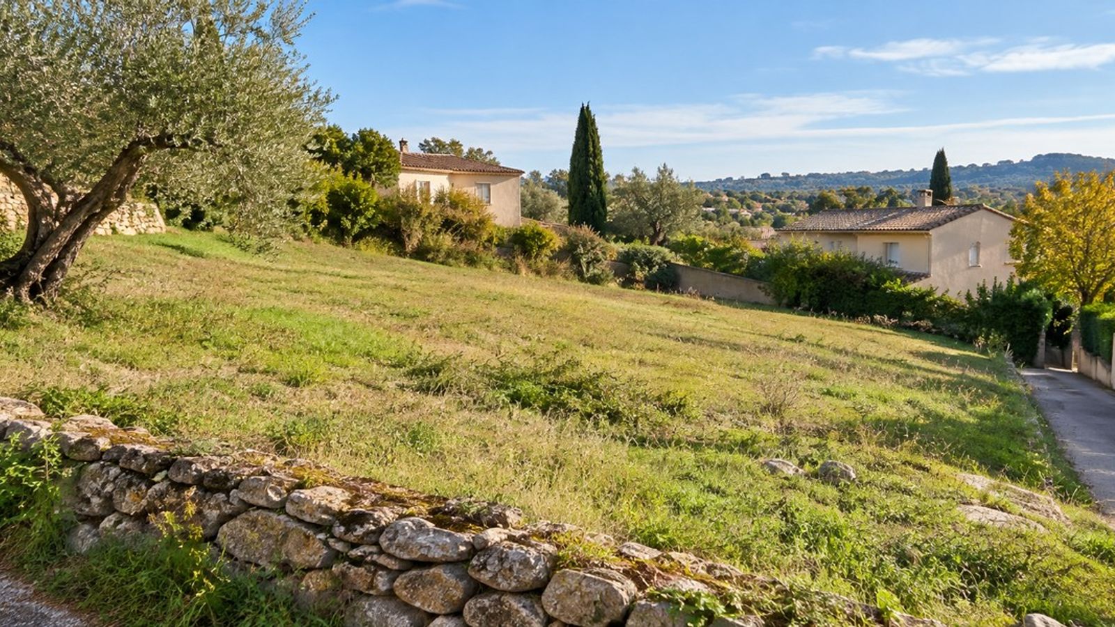 Terrain herbeux en pente douce, bordé d’un muret en pierre, avec une voie d’accès et des maisons en arrière-plan.