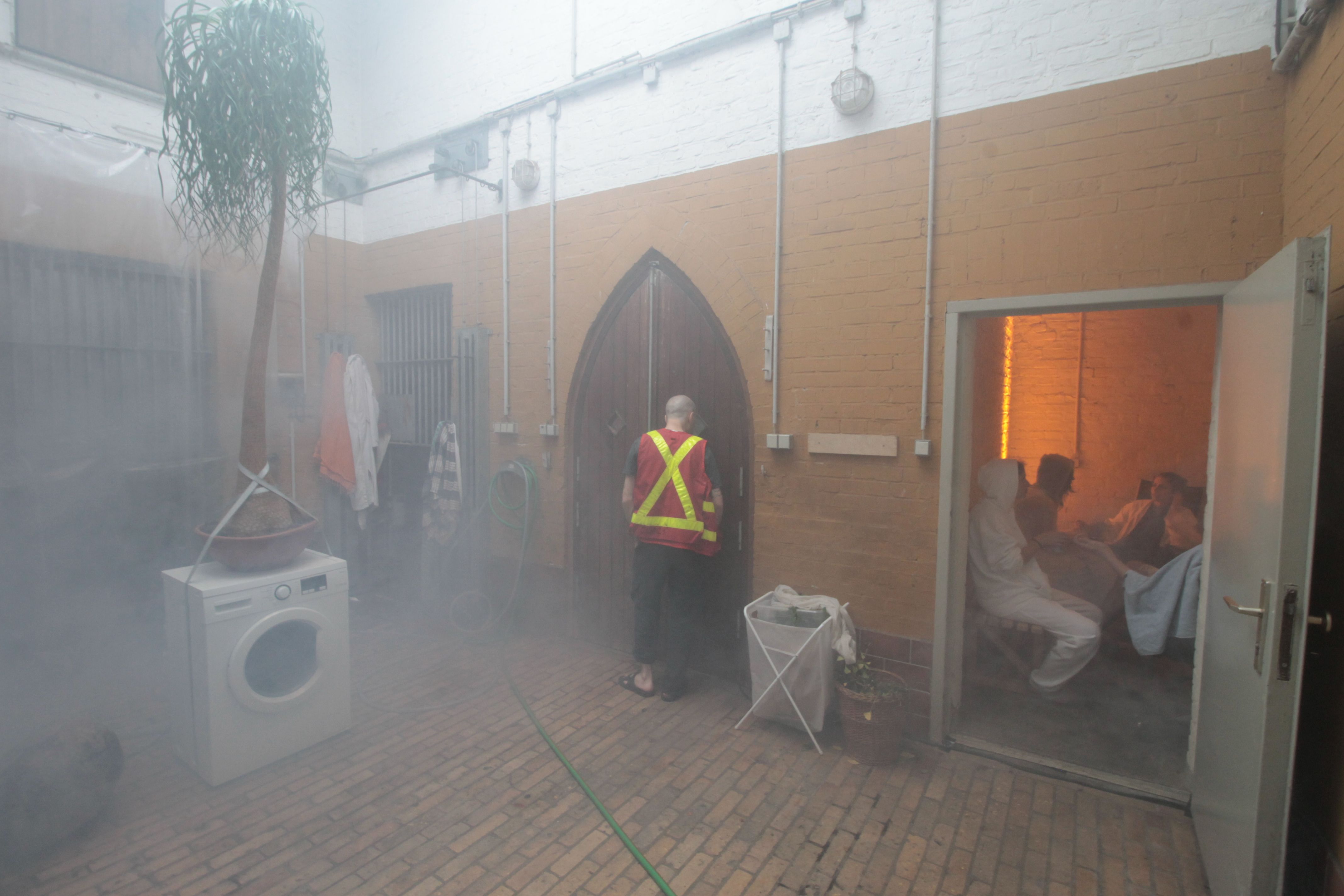 Installation view in bear cage of participants relaxing after sauna while technician working in central steamy room with herbal infusion washing machine in PPKK 03.00 sauna by Sarah Ancelle Schoenfeld and Louis-Philippe Scoufaras at Baerenzwinger Berlin