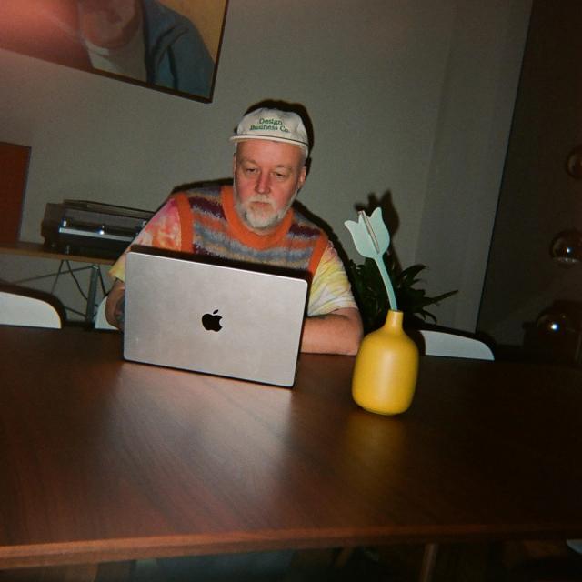Stewart Scott-Curran working on an MacBook Pro, with a green flower in a yellow pot nearby. In the background, a record player and a colorful painting.