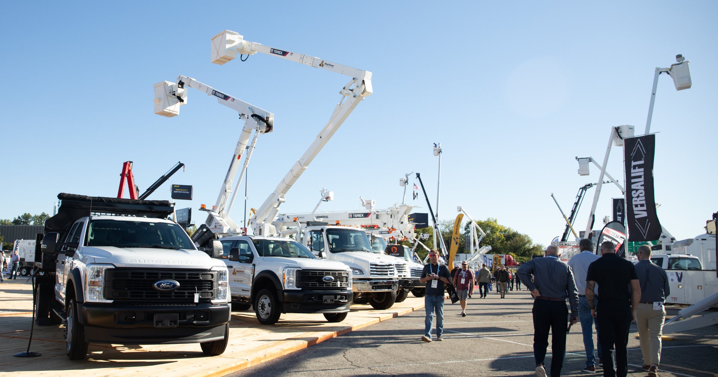 Utility fleet trucks and bucket trucks on display at an outdoor equipment expo, with attendees walking through the demonstration area under clear skies.
