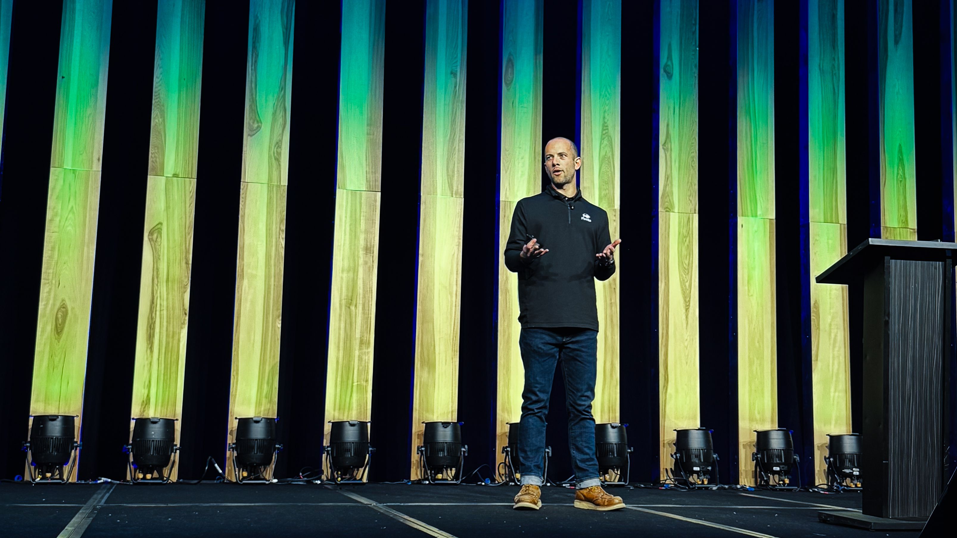 Speaker presenting on stage at a fleet technology conference, standing in front of tall illuminated panels with green and blue lighting.
