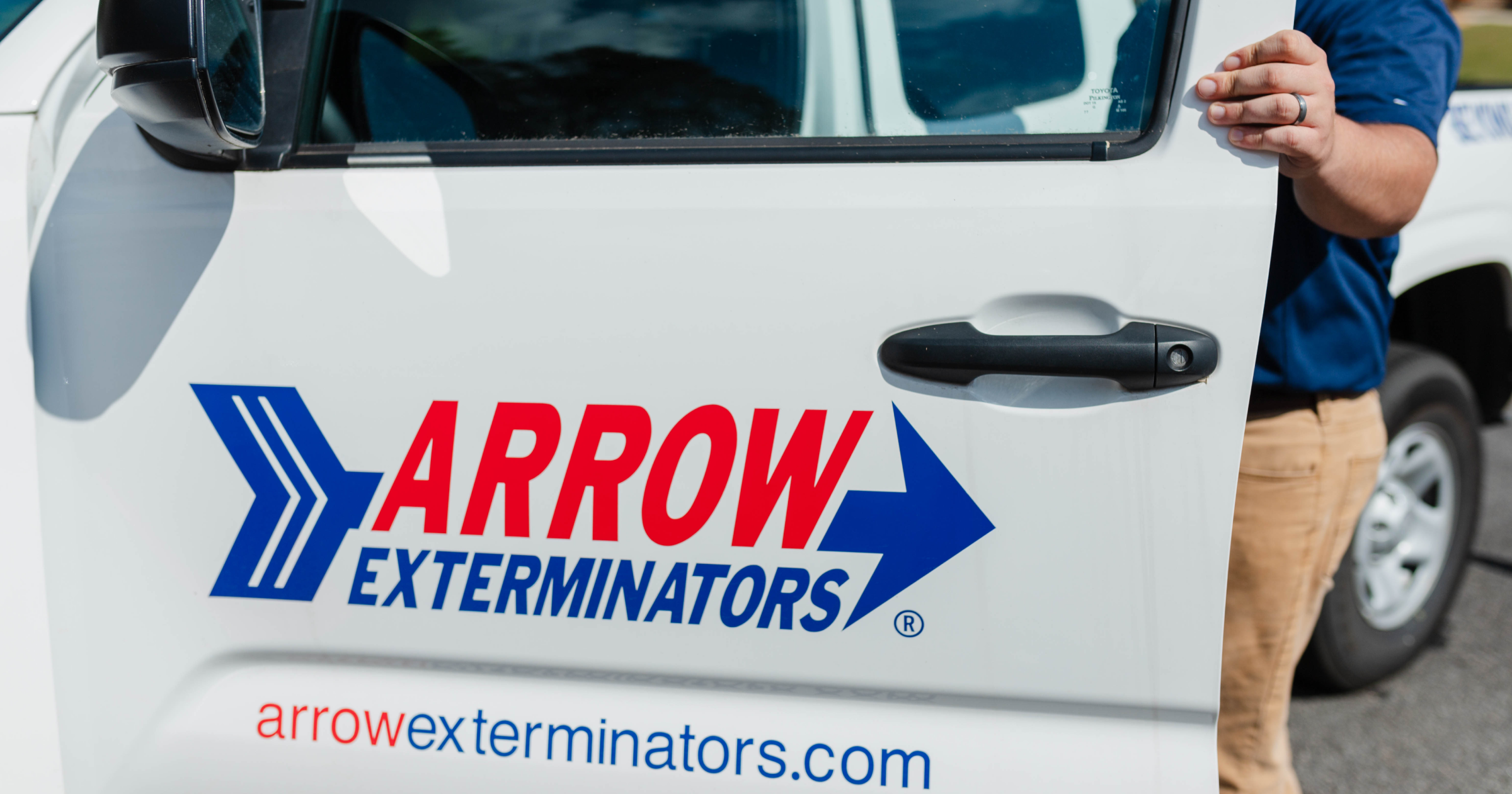 Close-up of an Arrow Exterminators service vehicle door with the Arrow Exterminators logo and website, with a technician standing beside the open door.