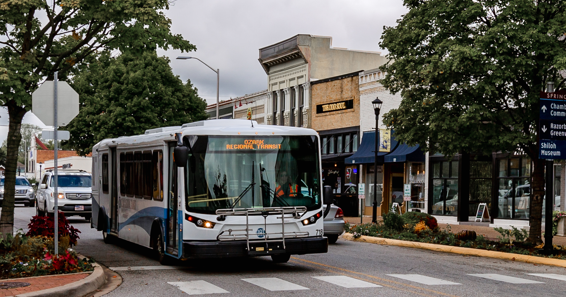 Ozark Regional Transit bus driving through a downtown street lined with shops, trees and parked cars