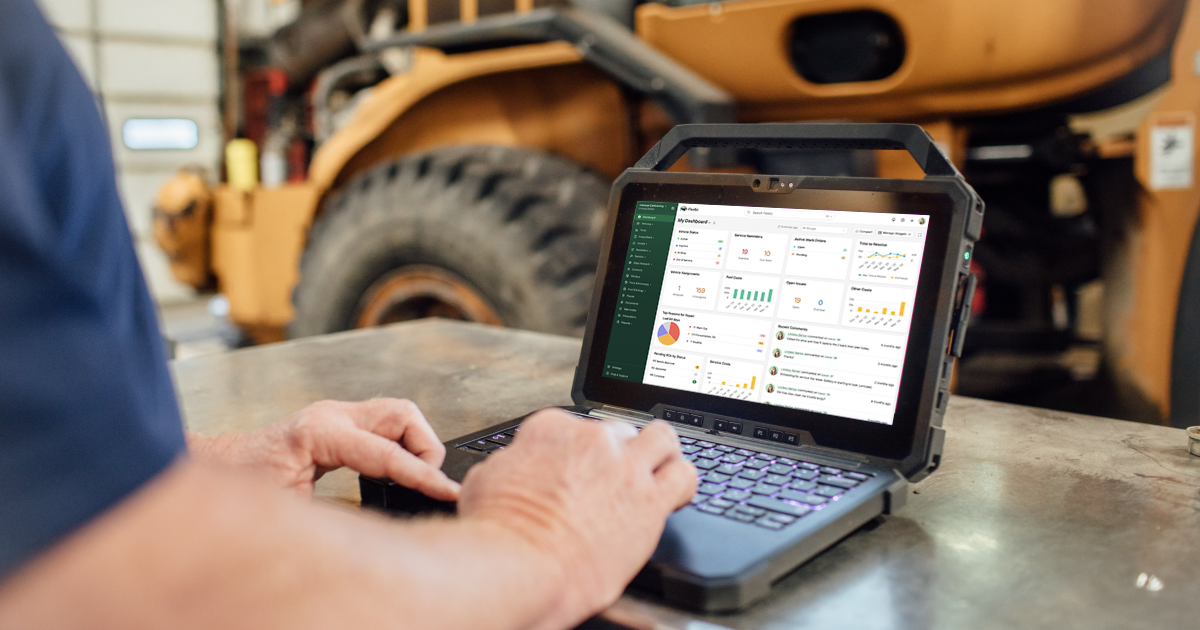 A heavy equipment technician uses a rugged laptop running Fleetio’s maintenance dashboard in front of a large wheel loader inside a service bay.