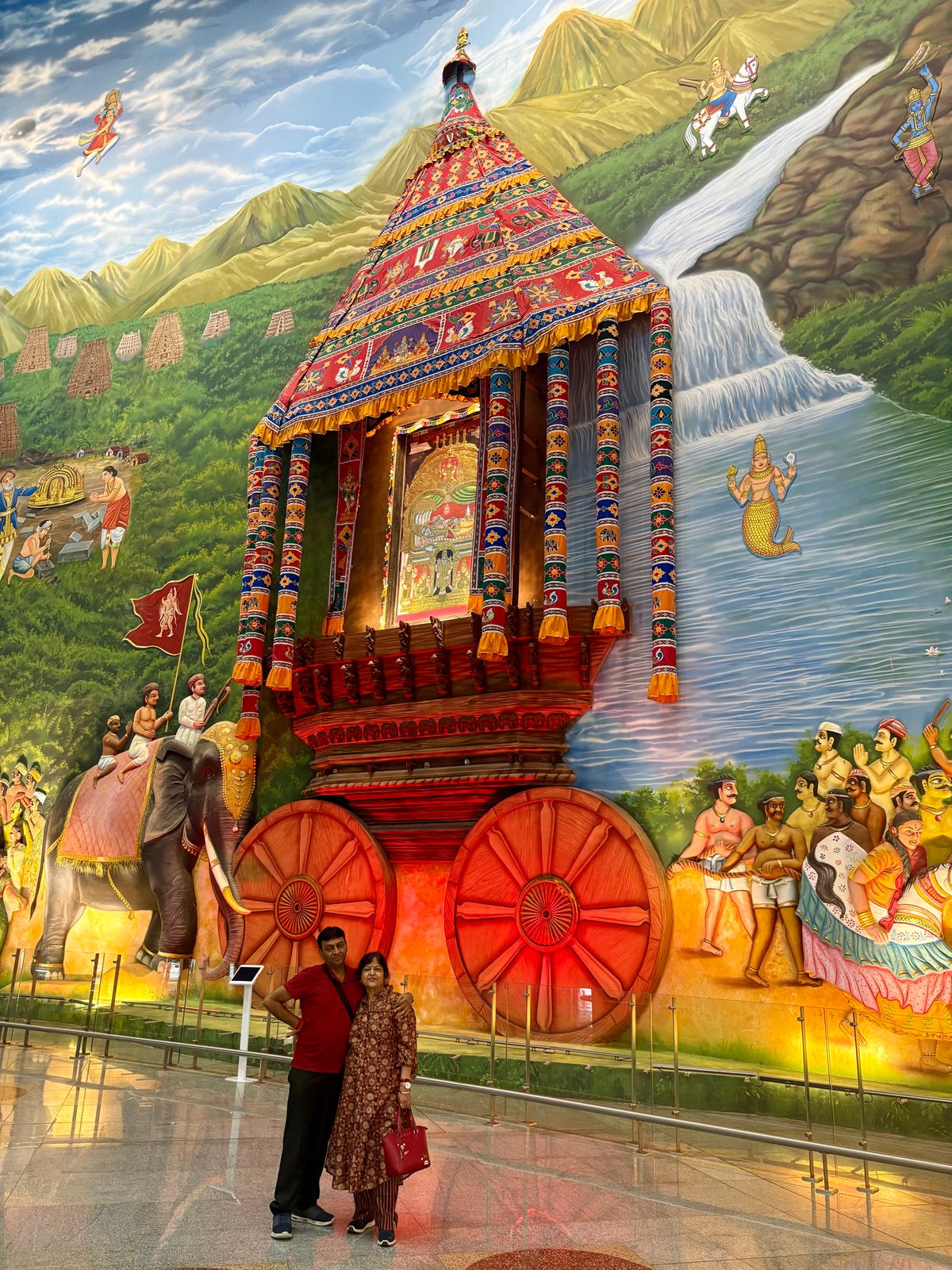 Ma and Papa at Tiruchirappalli airport — in front of a magnificent temple chariot (ther) installation set against a sweeping mural of Tamil Nadu's sacred heritage, a fitting farewell after the pilgrimage through the south.