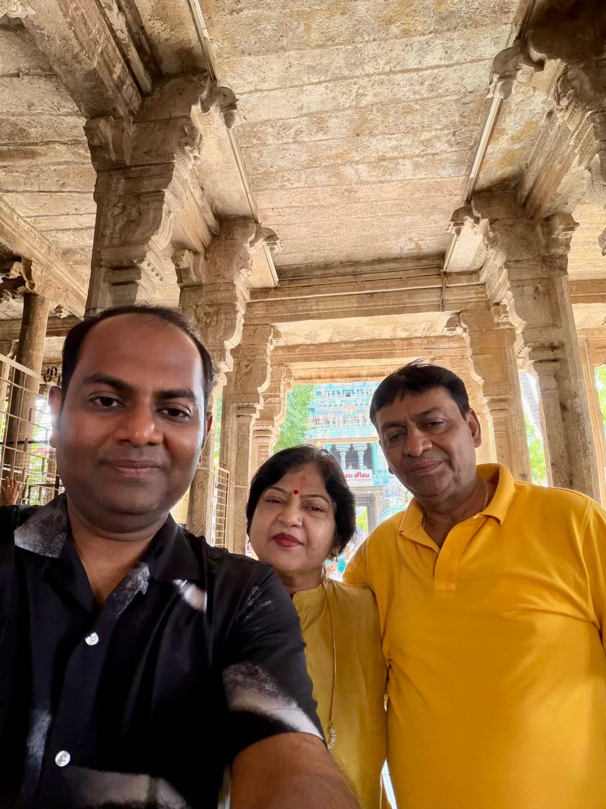 A selfie under the carved stone ceiling of Jambukeswarar Temple — the gopuram peeks through the columns behind. The three of them, barefoot on ancient stone, the sacred tank somewhere close.