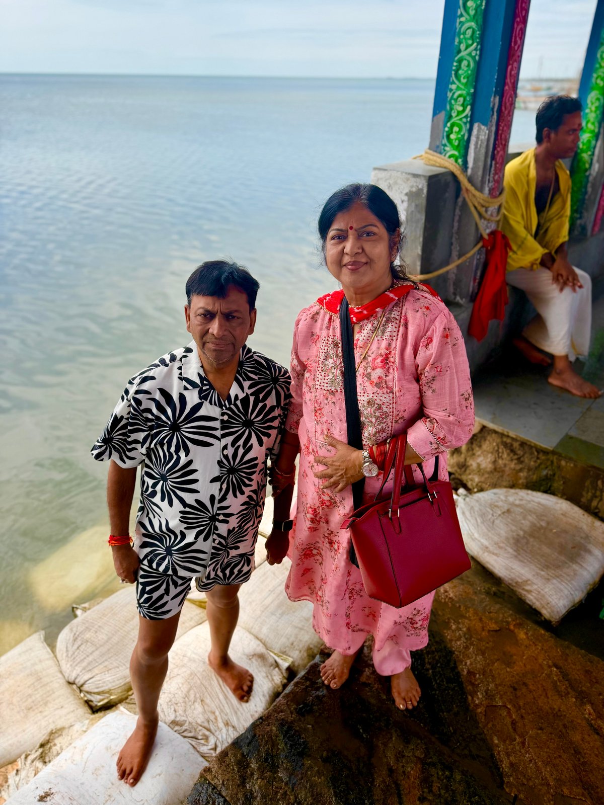 Standing at the water's edge at Devipattinam — the Palk Strait stretching to the horizon, the submerged Navagraha stones just below the surface. A priest rests nearby. This is one of the few temples in India where the deities are in the sea, not in a sanctum.