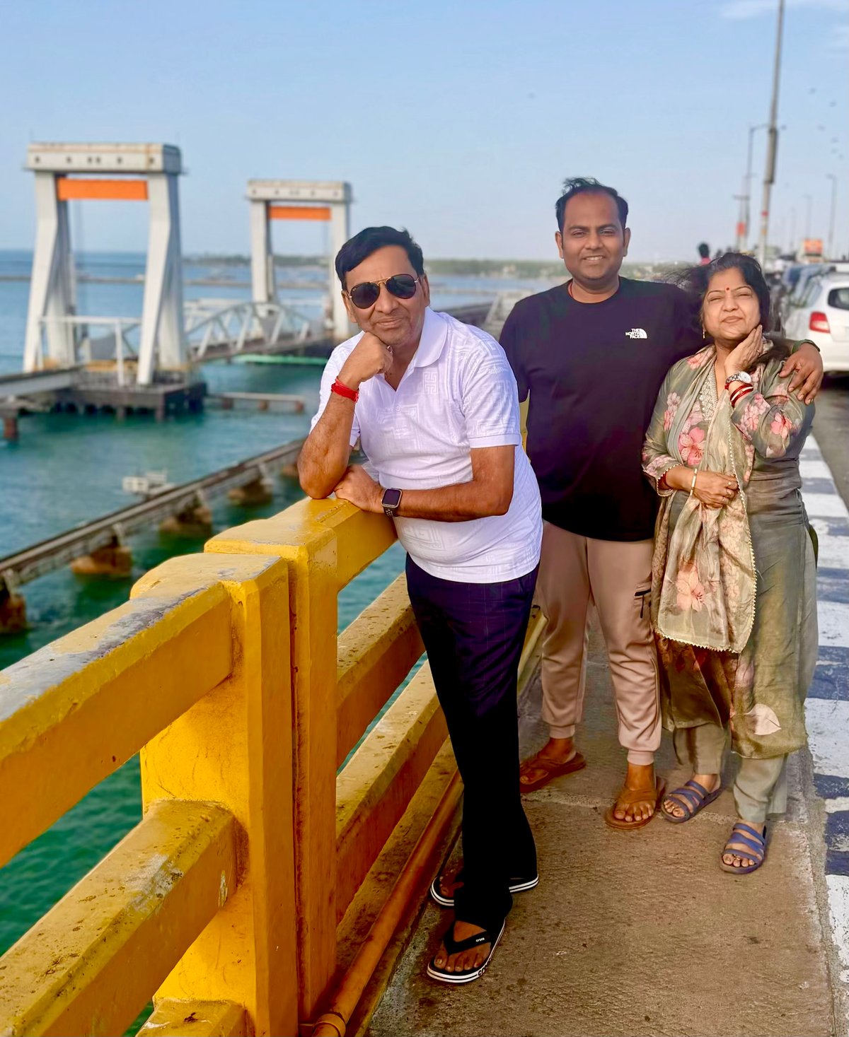 Papa, Shiv, and Ma on the Pamban Bridge — the iconic yellow railing, the open sea, and the railway bridge running parallel in the distance. One of those stops that earns its place on every itinerary.