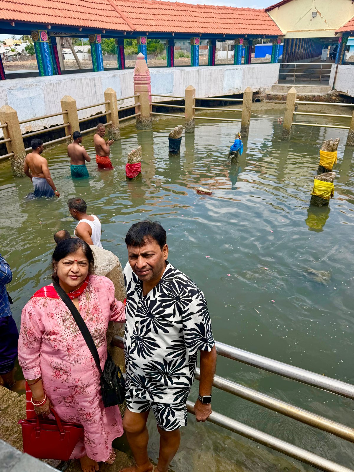 At the Navagraha kund — the sacred pool where devotees wade among the nine planetary stones. Behind Megha and Shiv, men are already in the water, circling the pillars in prayer. Each stone represents one of the nine planets of Vedic astrology. You go to each, barefoot in the water, and offer what you owe.