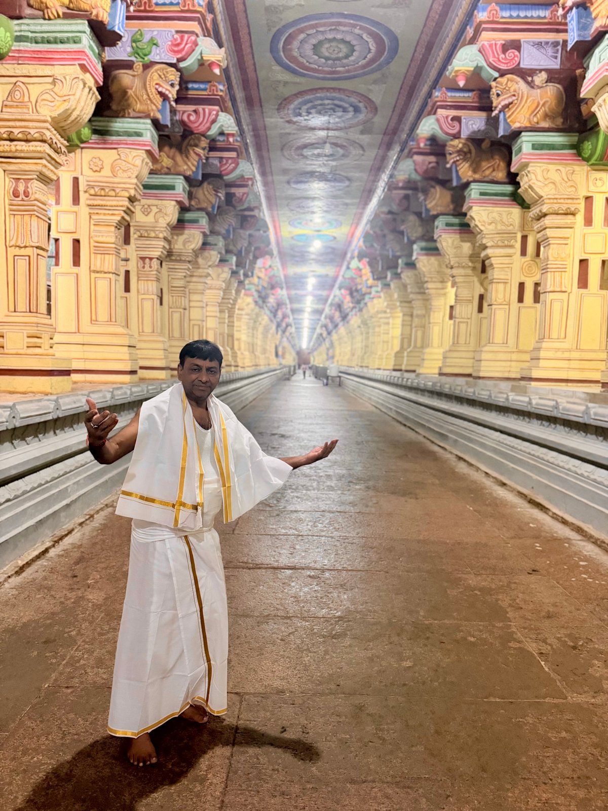 Standing in the world's longest temple corridor — Ramanathaswamy's third prakara runs 197 metres, lined with 1212 granite pillars on each side. The painted ceiling, the symmetry, the light at the far end: it doesn't look real. Shiv in traditional dhoti, arms open to it all.