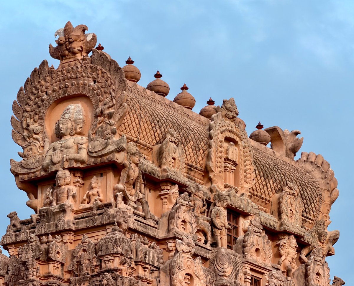 The crown of a gopuram — a four-faced deity in the topmost niche, flanked by celestial figures, the barrel-vaulted shikhara above them. At this level of detail, it stops being architecture and becomes scripture in stone.