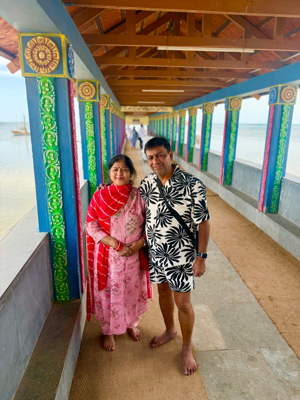 Ma and Papa in the covered walkway of Devipattinam Navagraha Temple — colourful pillars on one side, the Palk Strait on the other. Bright, breezy, and unlike any other temple on this trip.