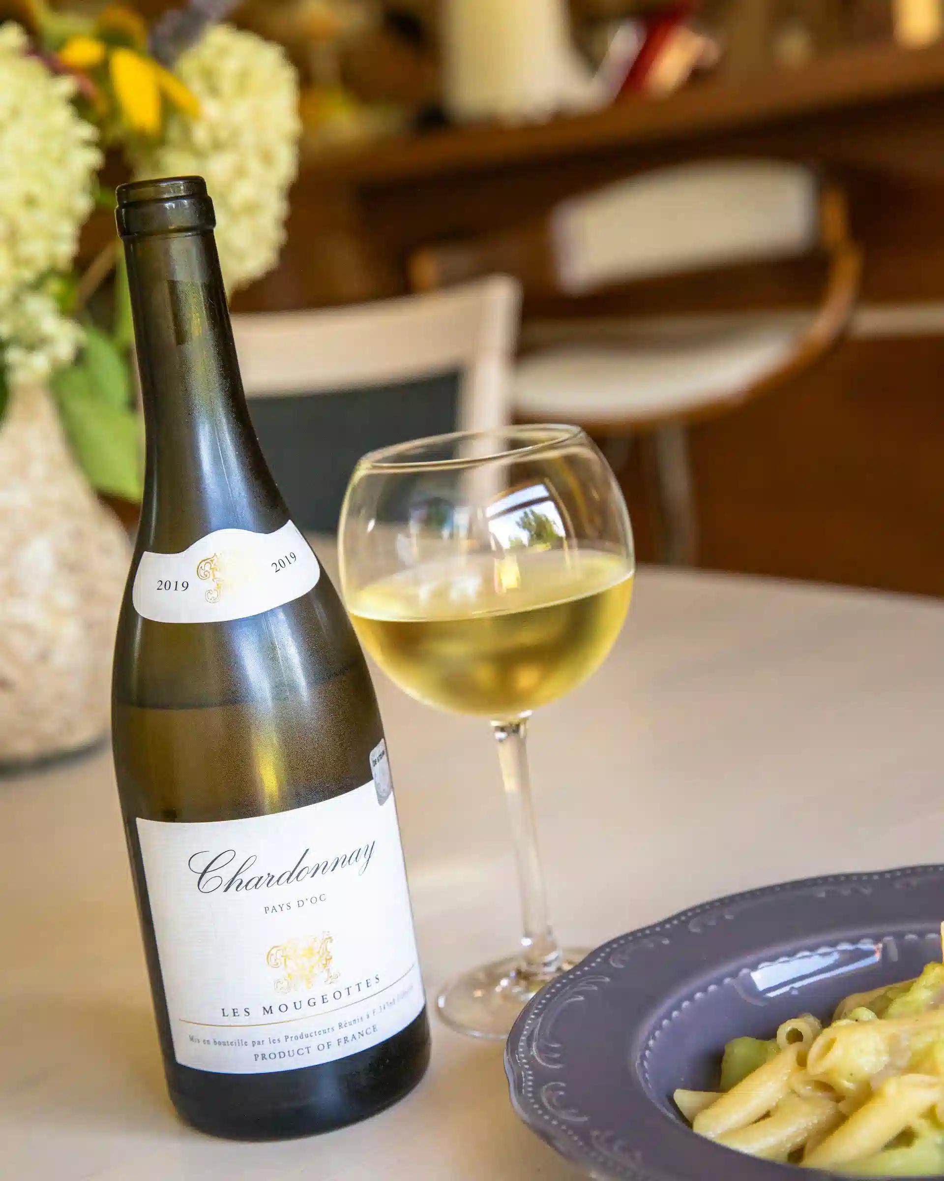 A bottle and glass of chardonnay with a glass of pasta on a table