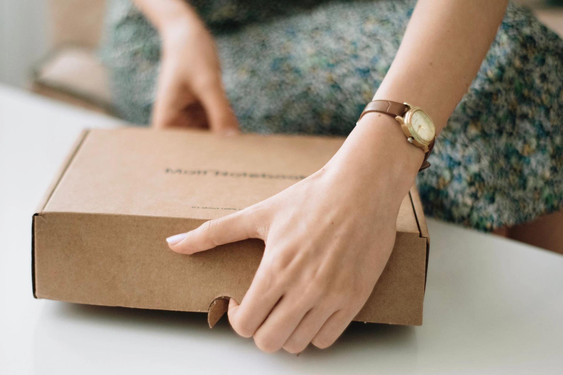 A woman opening a cardboard box with a watch-clad hand on a white table