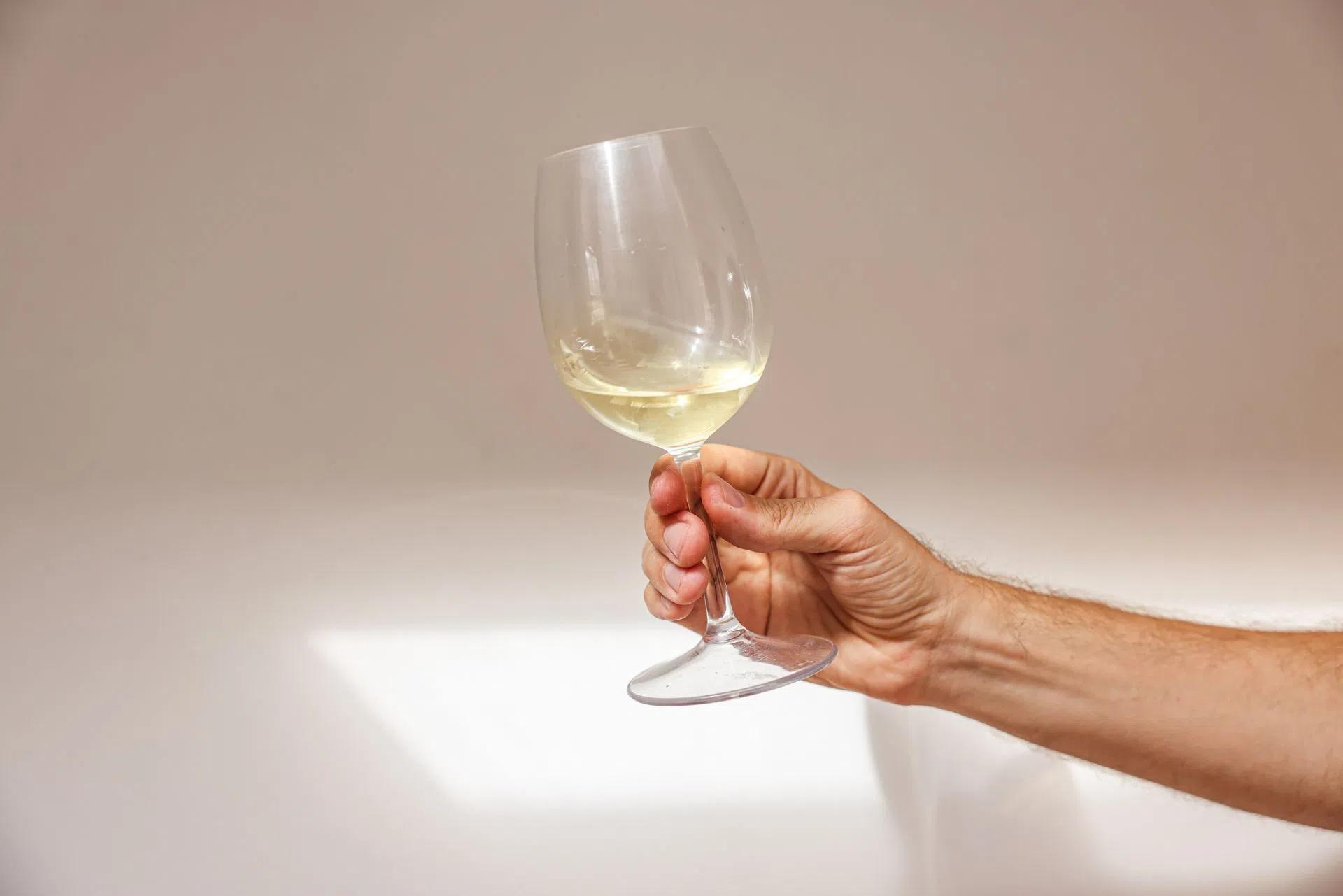 A close-up of a hand holding a glass of white wine against a plain white background, emphasizing elegance