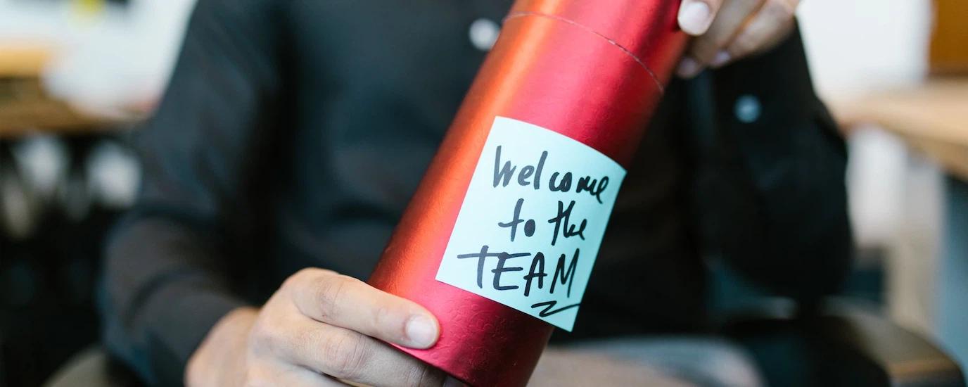 A red gift box held by a person with a welcome to the team note, ideal for new hire announcements