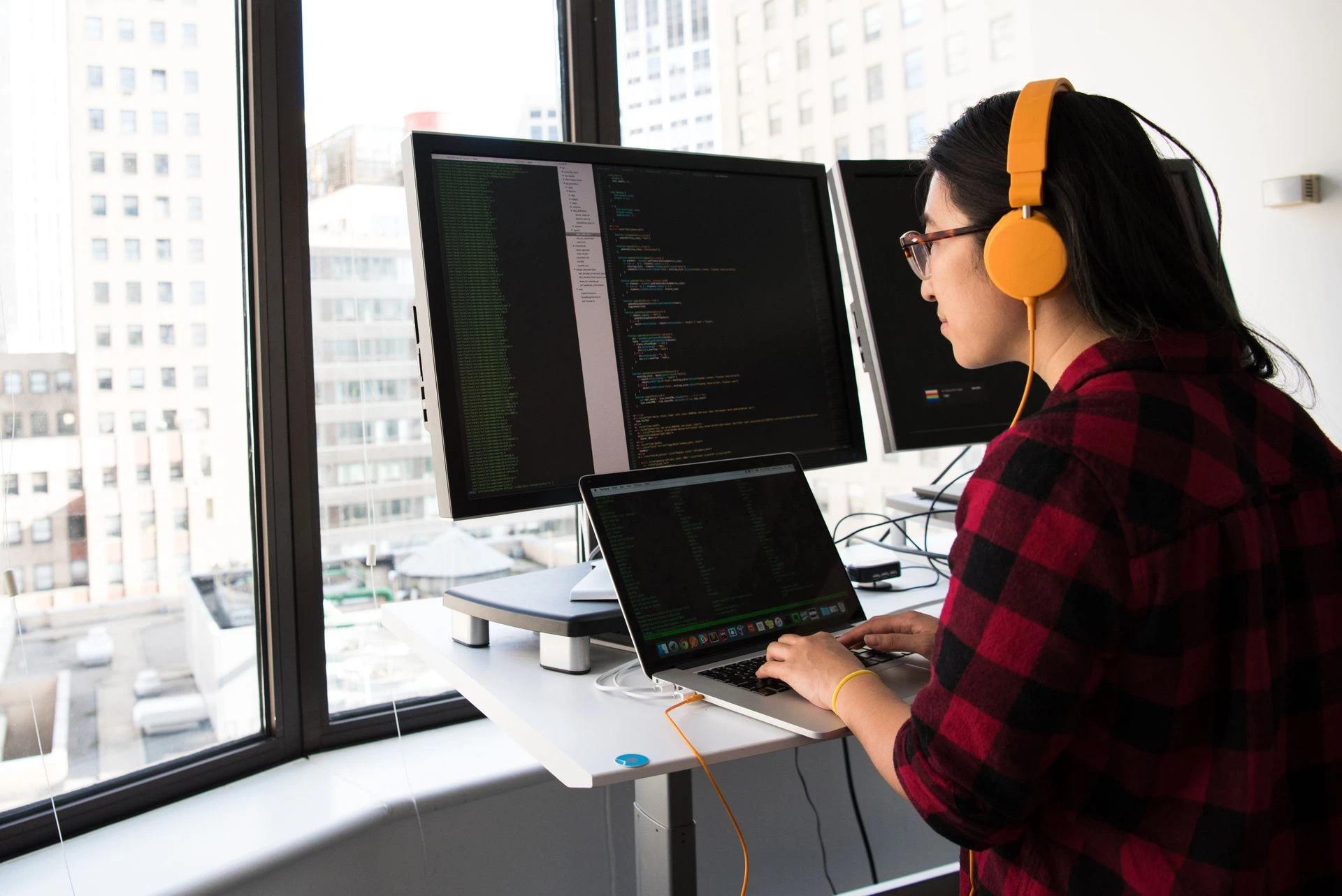Woman programming on a laptop at a standing desk in an office with large windows