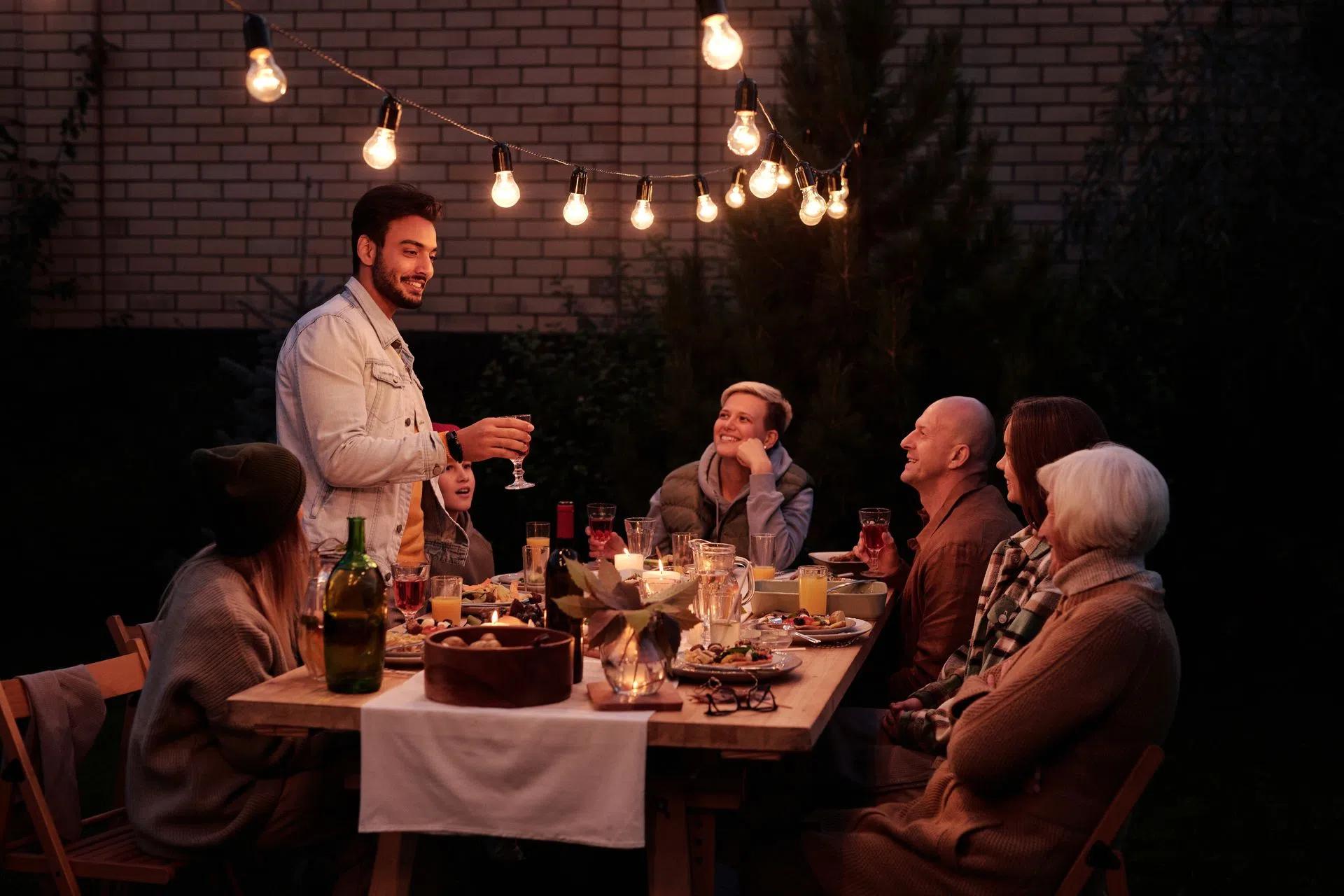 A group of adults enjoy a dinner party outdoors under warm string lights, sharing food and smiles.