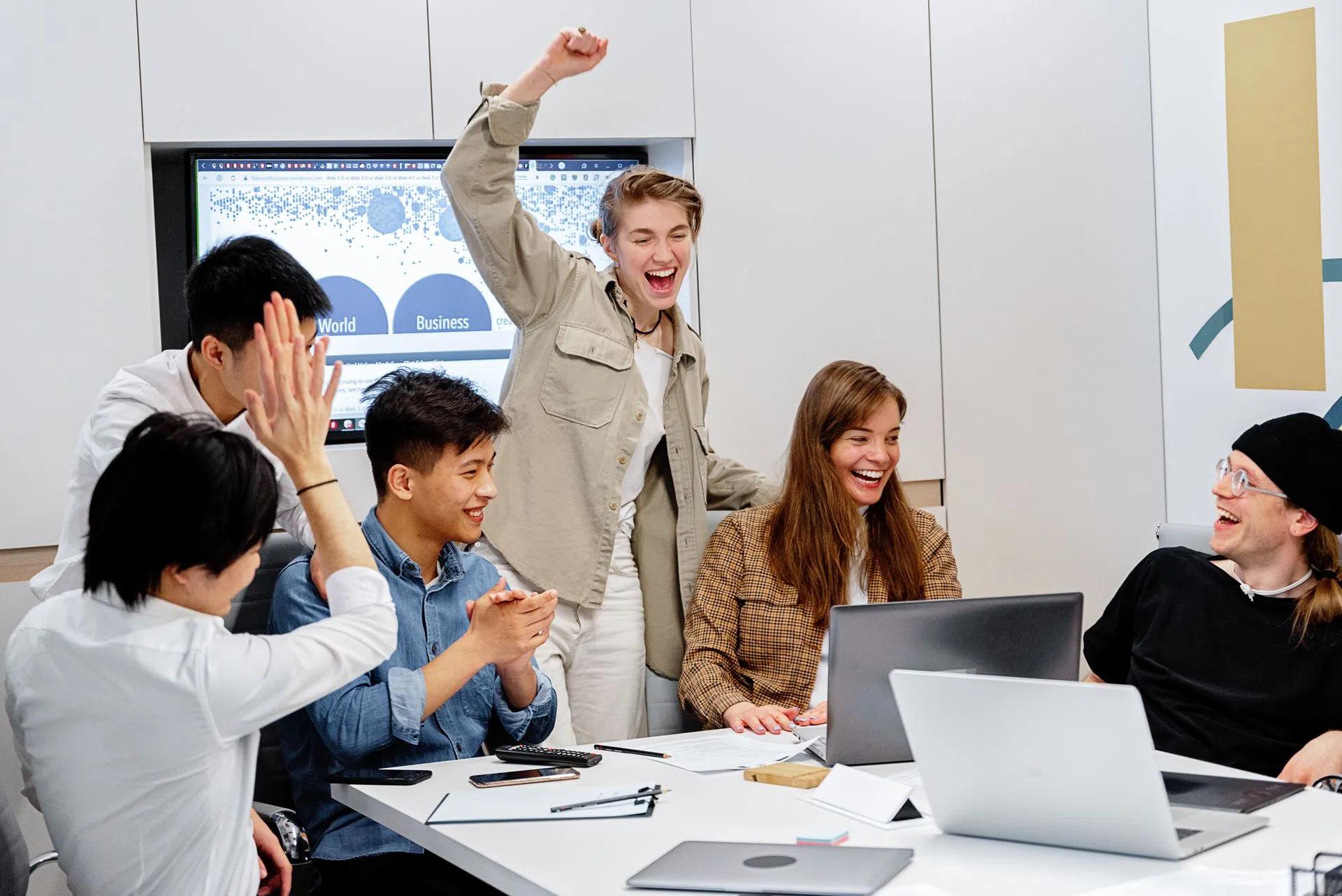 A diverse team enthusiastically celebrating success around laptops during an office meeting