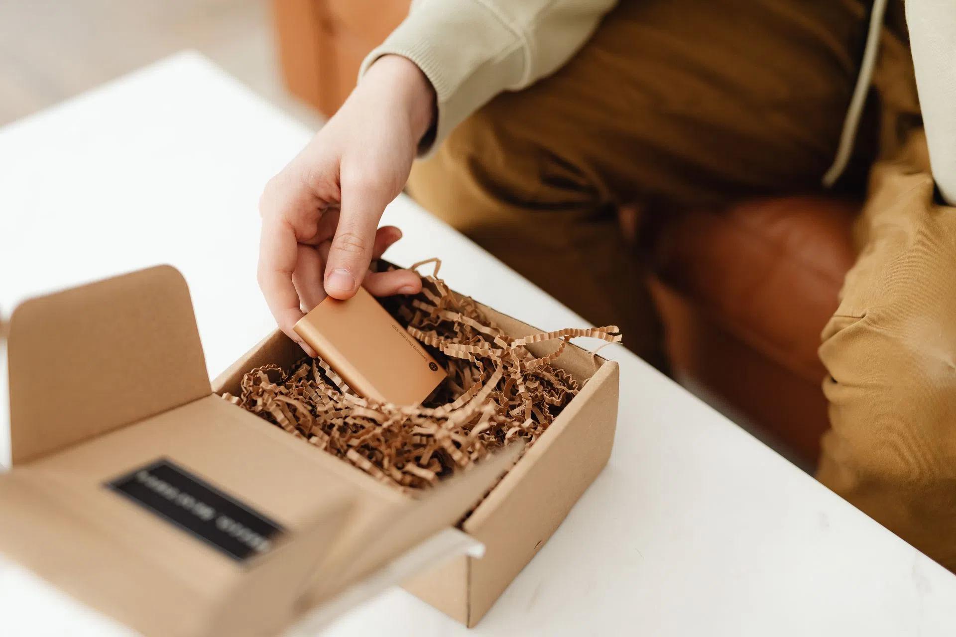 A person unboxes a small item from a cardboard package filled with crinkle paper, indoors