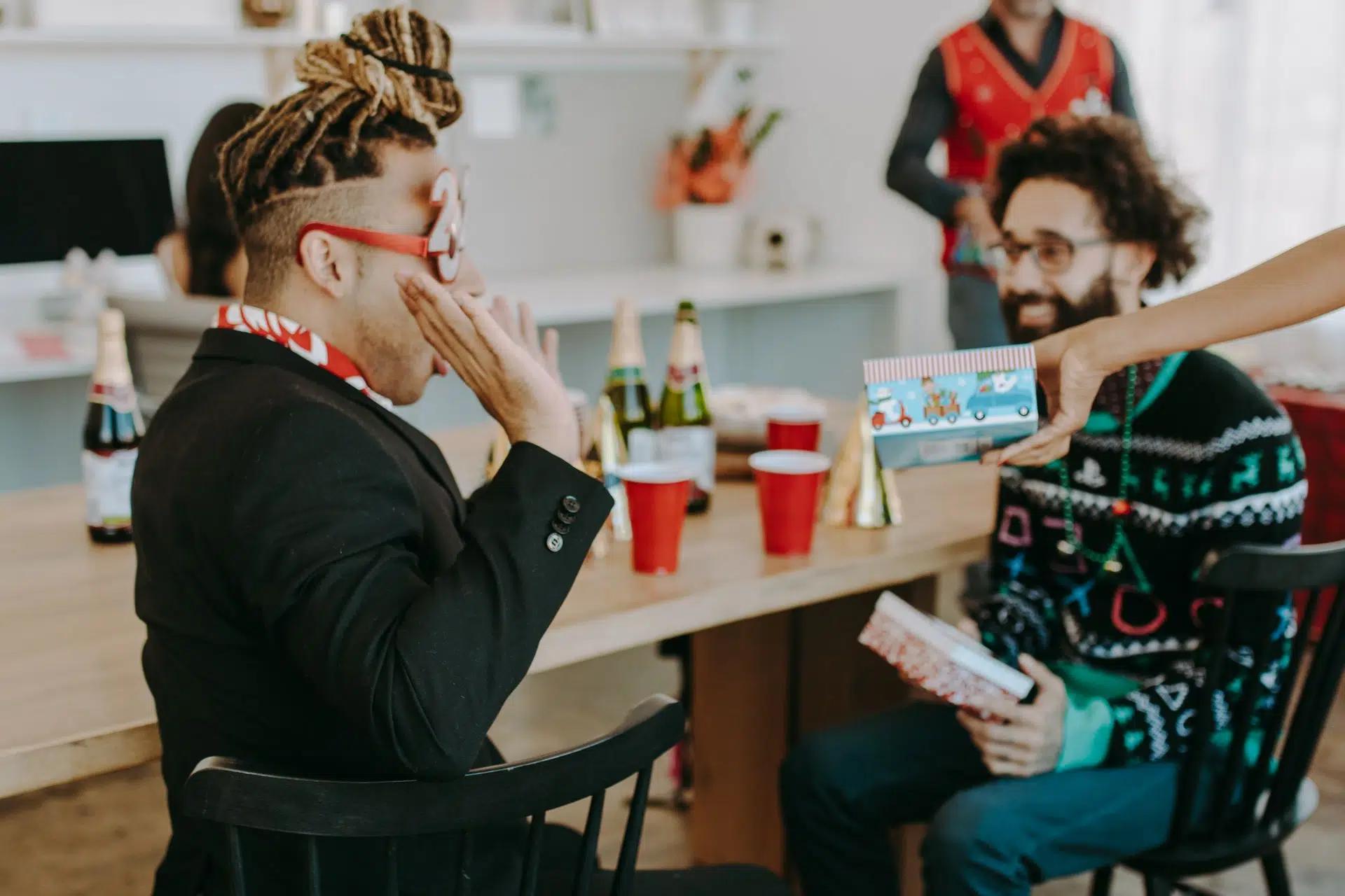 Group of adults enjoying a holiday party with festive sweaters, gifts, and drinks indoors