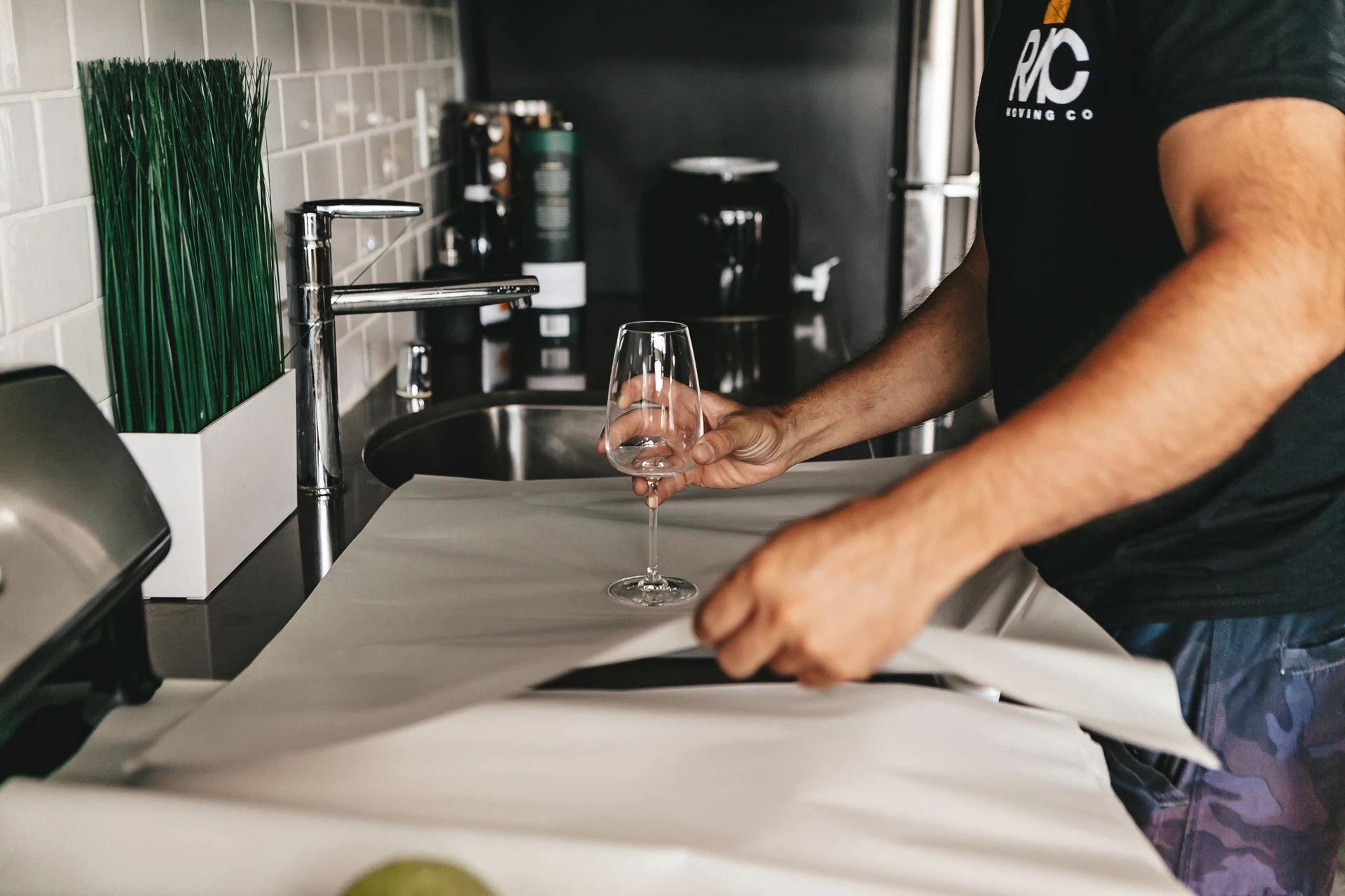 Person carefully wraps a wine glass in paper on a kitchen countertop, focusing on safe packing and organization.