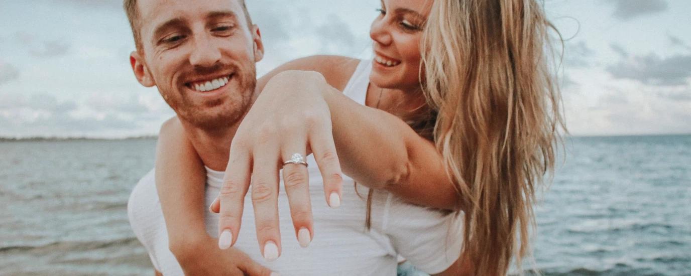 Happy couple embracing by the ocean, showcasing an engagement ring in a joyful moment