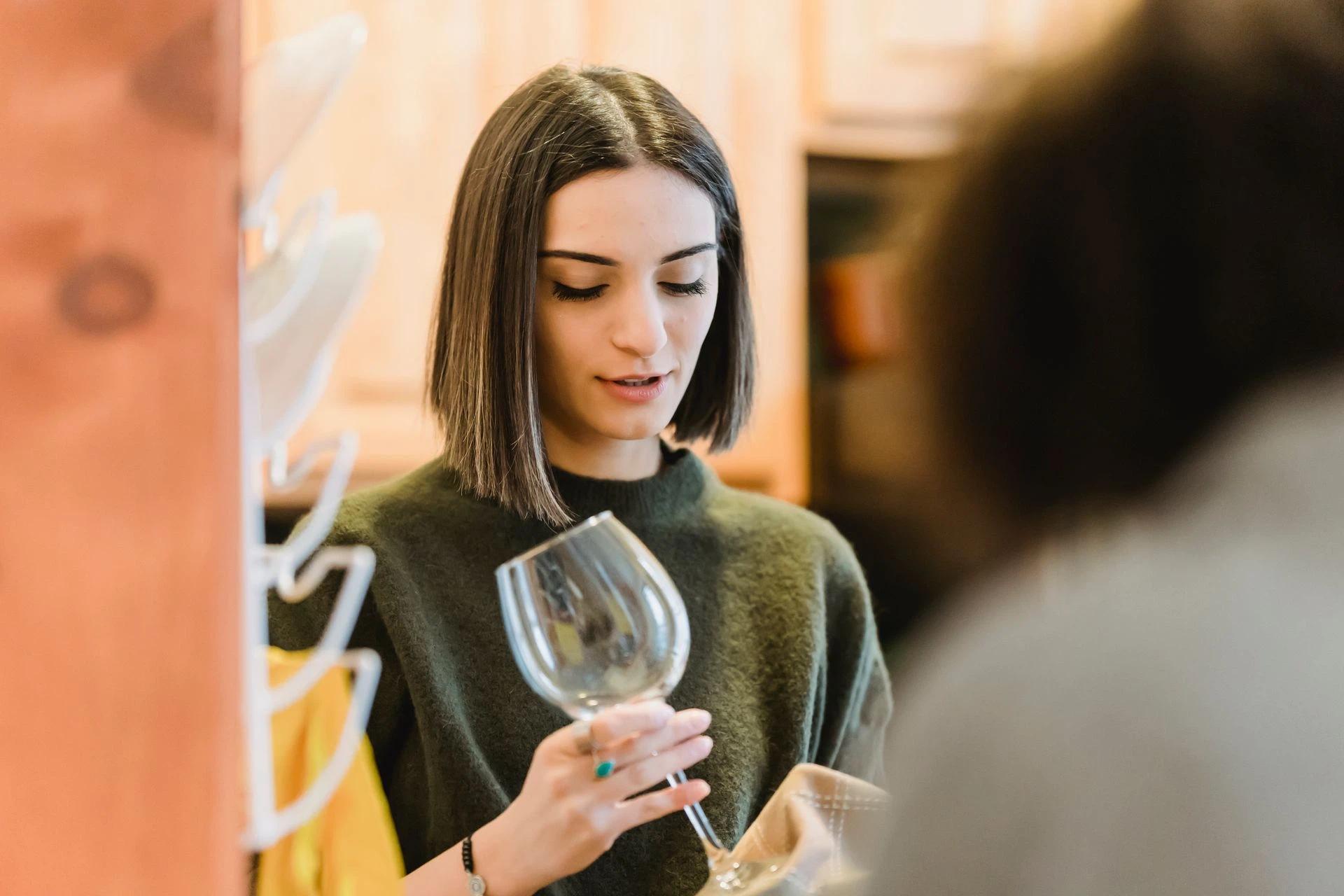 A young woman is drying a wine glass while chatting with a friend in a cozy kitchen.