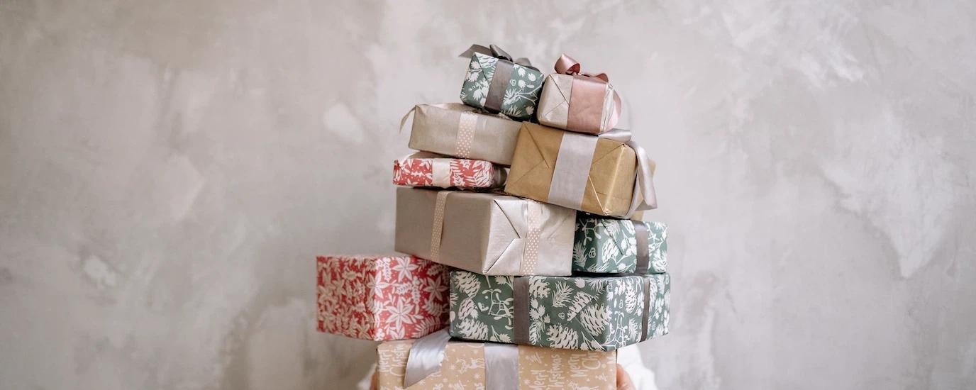 Person hiding behind a tall stack of gift-wrapped boxes against a gray wall, indoors