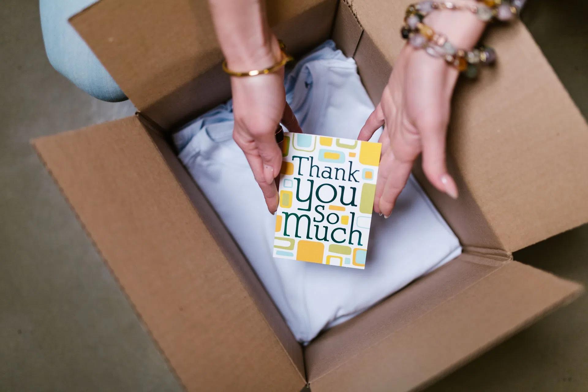 A person holds a thank you card above items in an open shipping box, suggesting care in sending