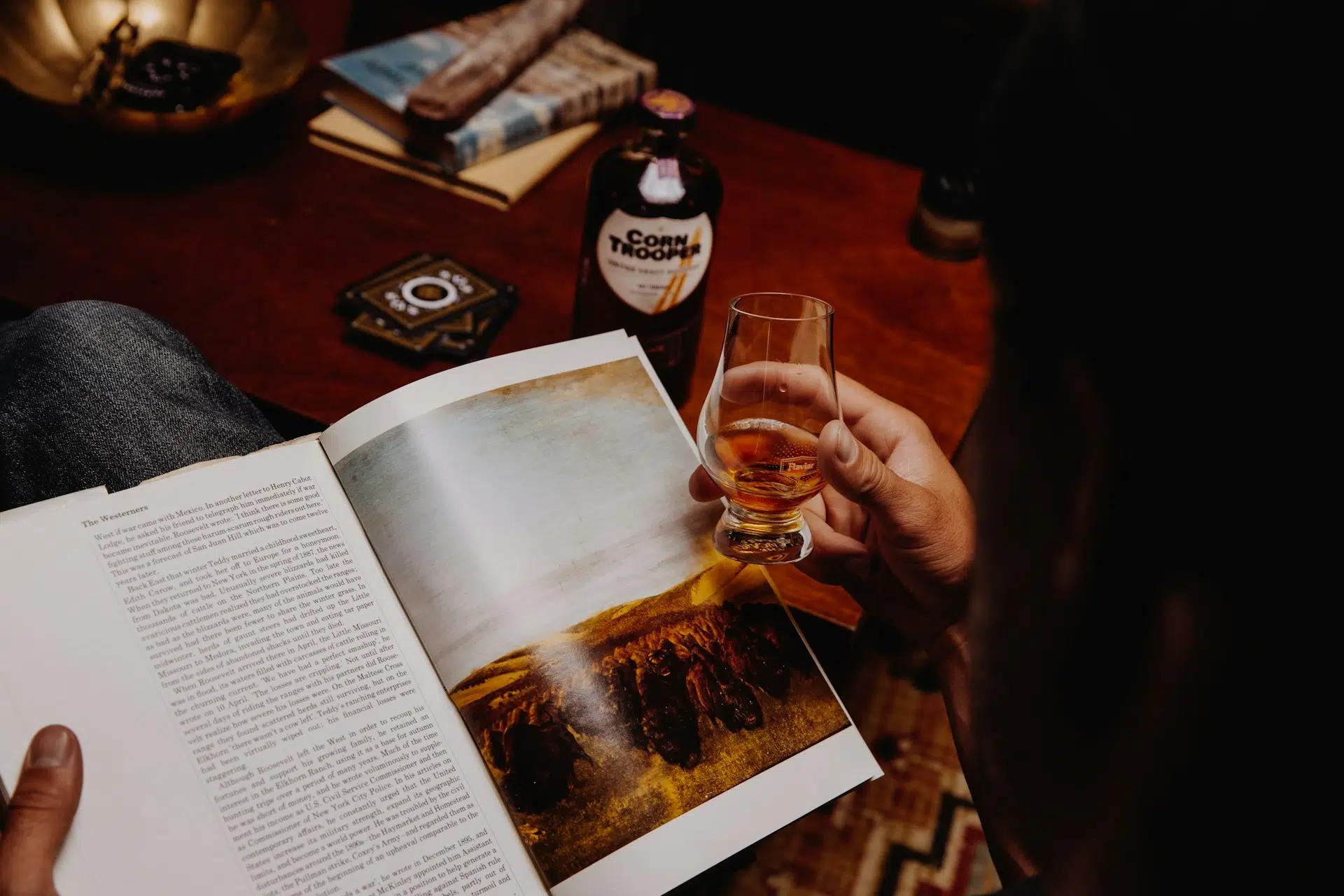 A person holding a whisky glass while reading a magazine
