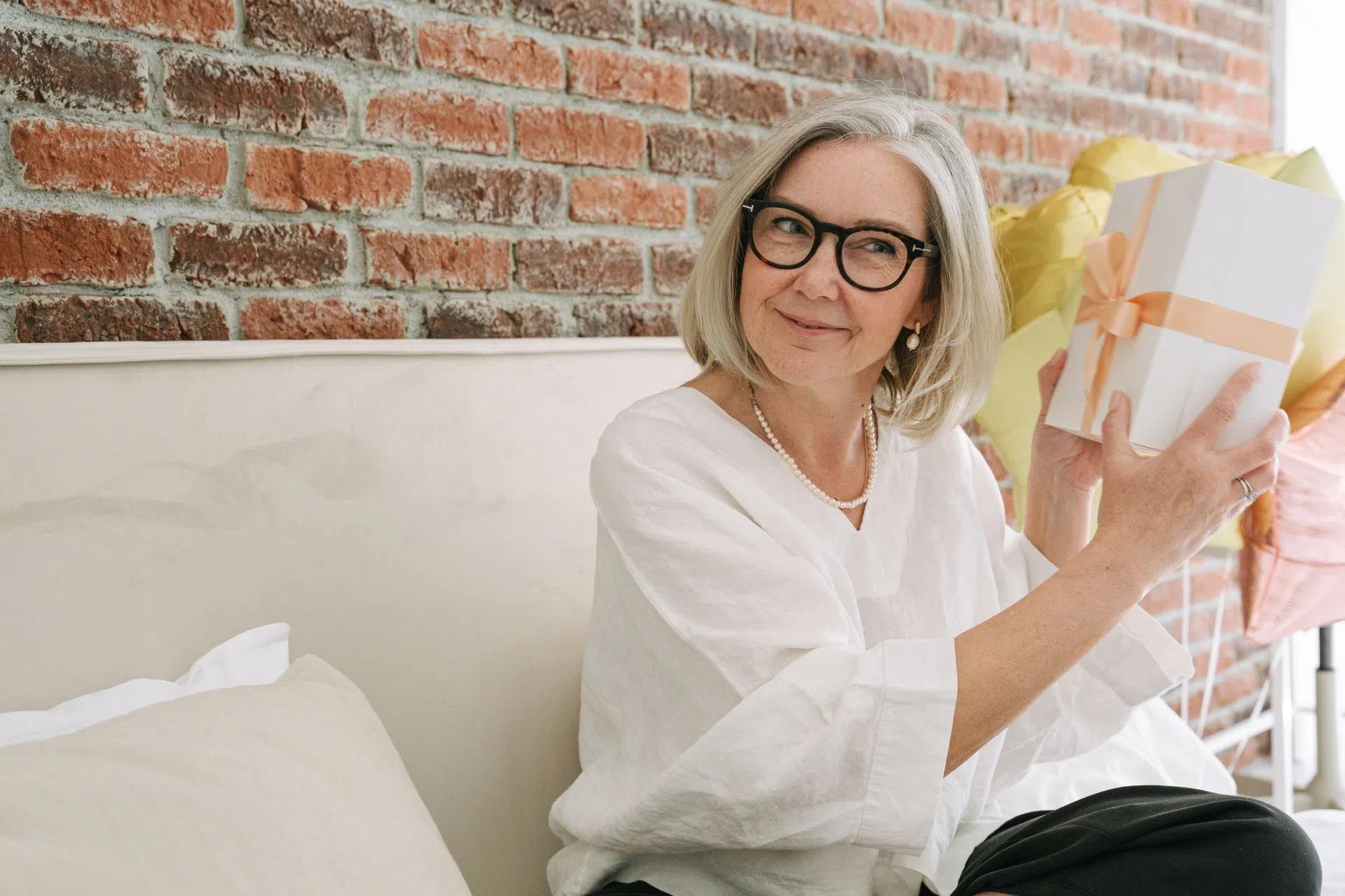 Senior woman in white blouse happily holding a gift box indoors in front of a brick wall