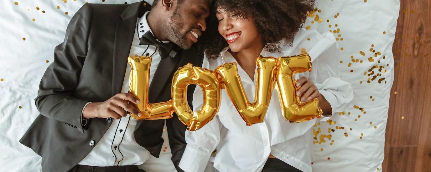 Smiling black couple lying on bed with LOVE balloons, joyful and intimate moment