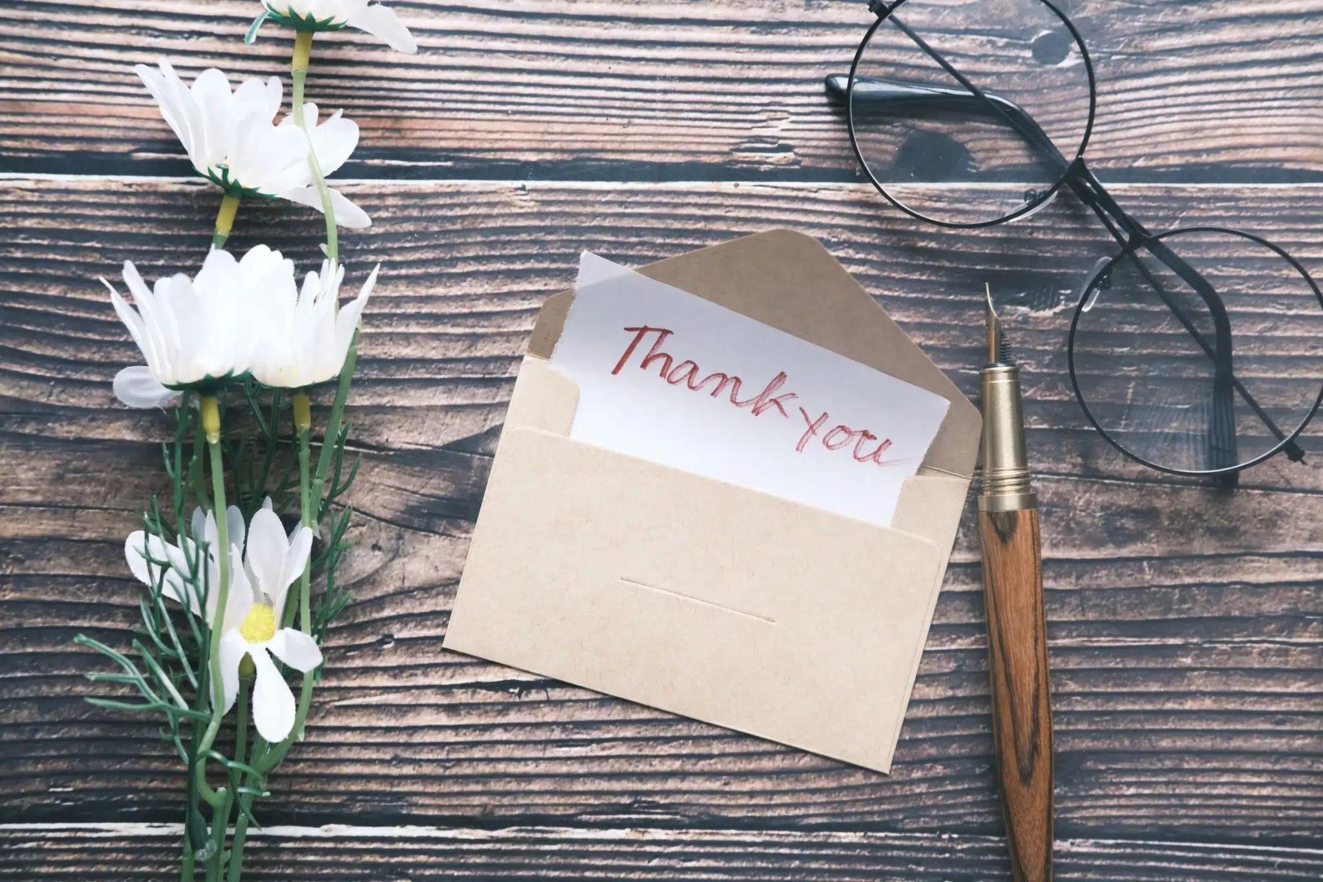 A flat lay of a thank you note in an envelope with daisies, glasses, and a pen on a wooden surface