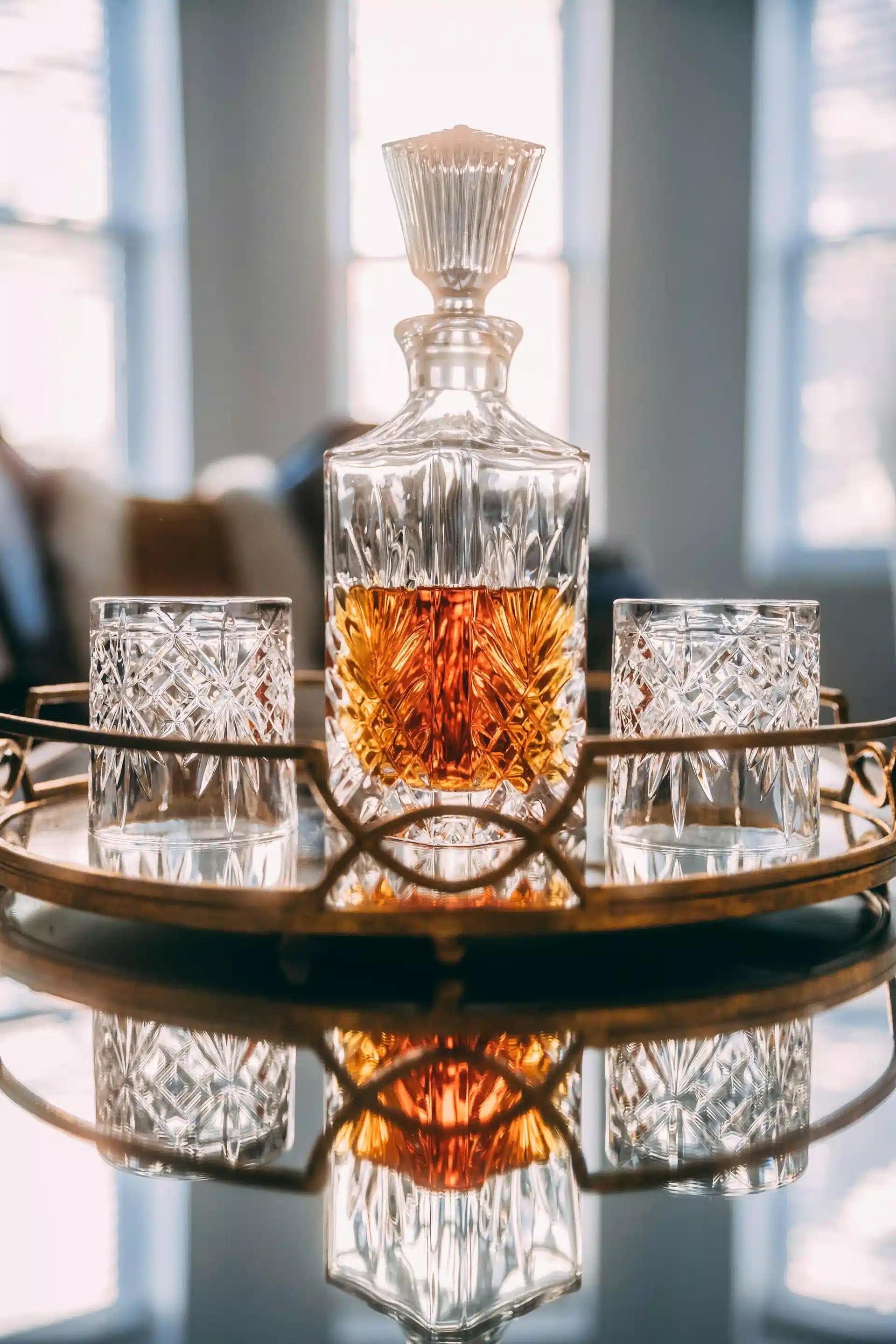 A filled up whiskey decanter with two empty whiskey glasses on a serving tray