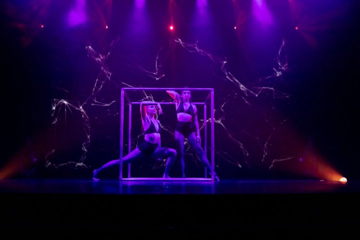 Twee vrouwelijke hedendaagse danseressen poseren in een grote metalen kubus op een podium van een cruiseschip, verlicht door dramatische paarse spotlights tegen een achtergrond met bliksemflitsen.