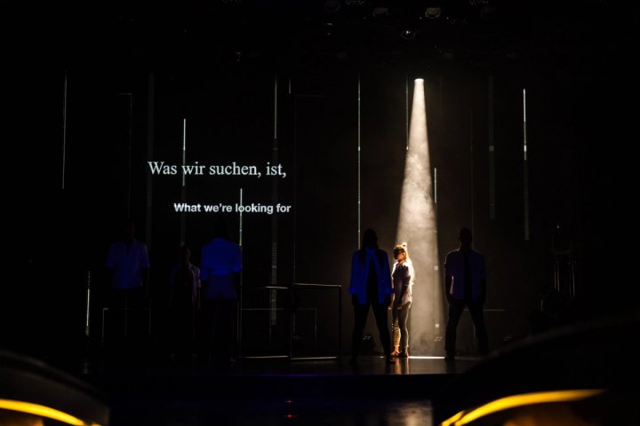 De castleden treden op in een dramatische, minimalistische productie op een cruiseschip, onder een schijnwerper, met de tekst 'Was wir suchen, ist, Wat we zoeken' geprojecteerd op een donker podium.
