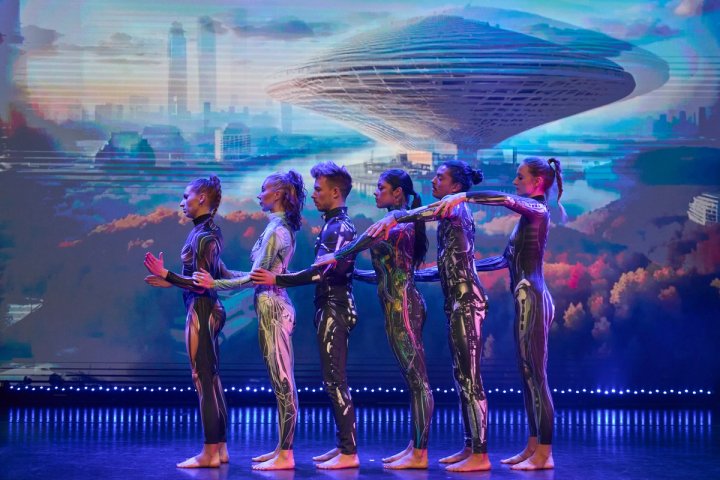 Een groep artiesten in futuristische, bedrukte bodysuits staat in een rij op een podium van een cruiseschip, met op de achtergrond een futuristische stad en een groot, iconisch architectonisch bouwwerk.