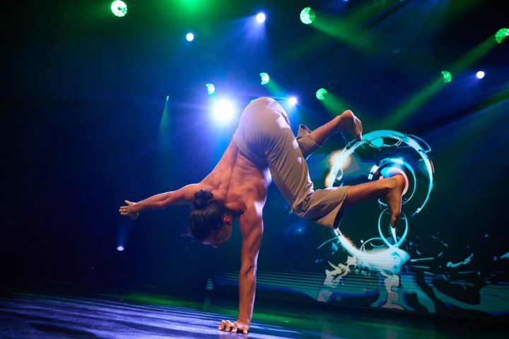 Een mannelijke hedendaagse danser voert een handstand op één hand uit op een podium van een cruiseschip, met dynamische groene en blauwe verlichting en een abstracte digitale achtergrond.