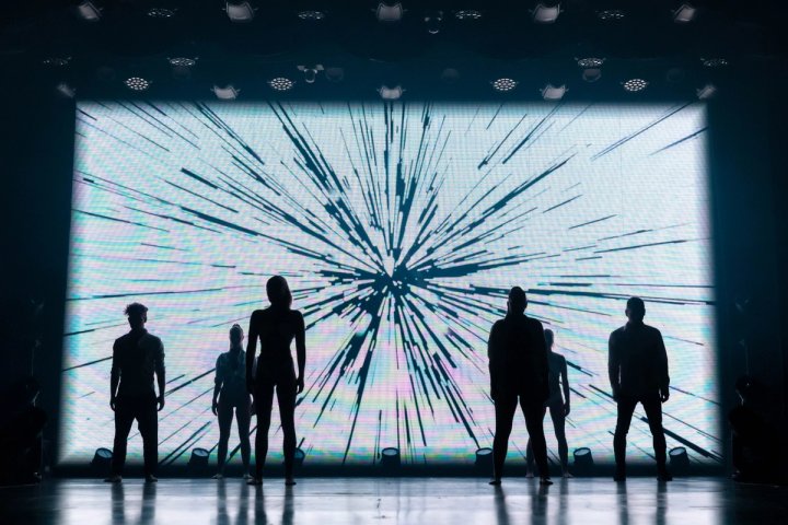 Castleden staan ​​in silhouet op een podium van een cruiseschip tegen een helder, exploderend digitaal scherm met radiale lichtstrepen tijdens een theatervoorstelling.