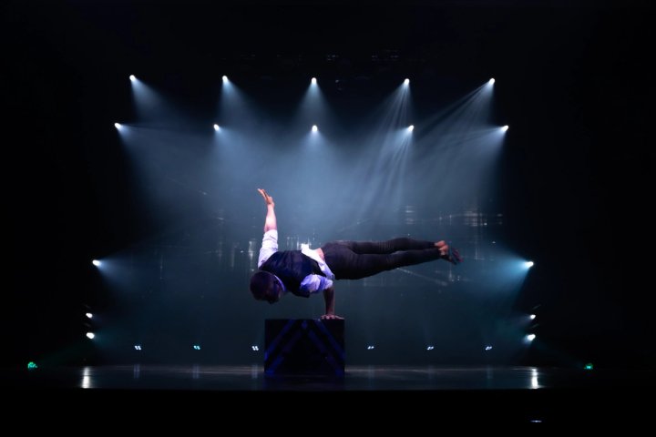 Een mannelijke acrobaat voert een krachtige planche-balansact met één hand uit op een platformblok, midden op het podium onder dramatische witte schijnwerpers tijdens een show op een cruiseschip.