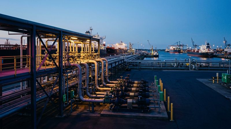 Mid-distance shot of pipeline terminal manifolds stretching toward a commercial shipping dock, warm industrial lighting highlighting heavy steel valves and transfer pumps under a calm evening sky.