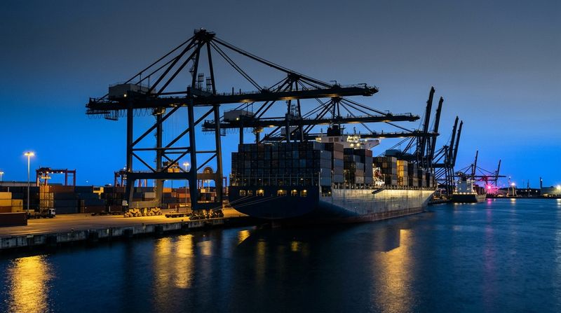 Deep-water commercial shipping port at dusk, rows of towering massive steel container cranes silhouetted against a fading sky, a colossal cargo ship being loaded with steel shipping containers, wide midshot framing, calm deep navy water reflecting warm yellow dockside illumination.
