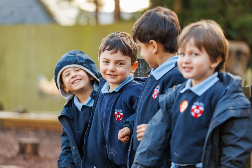 Four boys in navy blue uniforms at Mill Hill Pre-Prep smile and interact outdoors, enjoying a playful moment in the school playground.