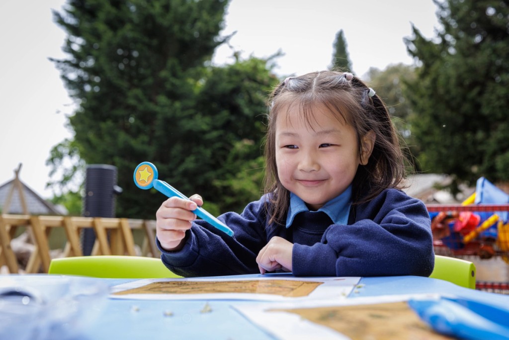 Young girl at Mill Hill Pre-Prep engaged in an outdoor learning activity, holding a pen with a star topper, smiling while sitting at a table with colourful educational materials.