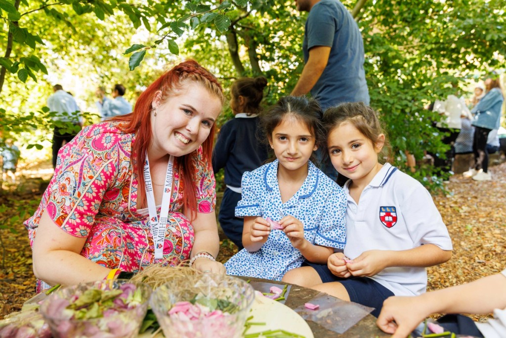 Teacher and two young girls at Mill Hill Pre-Prep engaged in an outdoor crafting activity, smiling together while surrounded by nature, creating a hands-on and interactive learning experience.