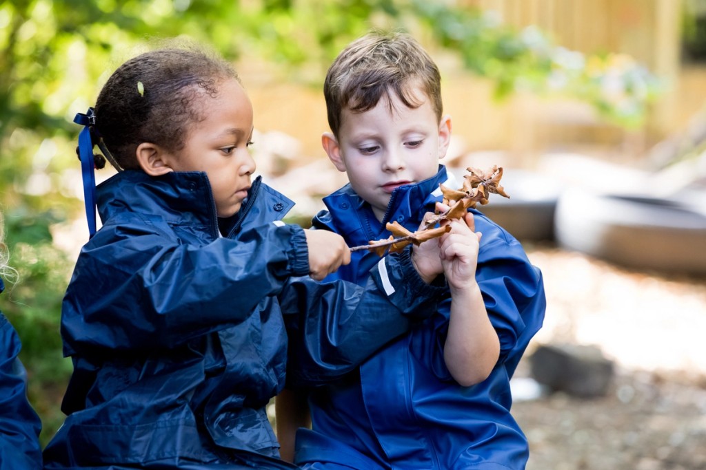 Two children in blue waterproof jackets sit outdoors at Mill Hill Pre-Prep, carefully examining autumn leaves together during a nature activity.